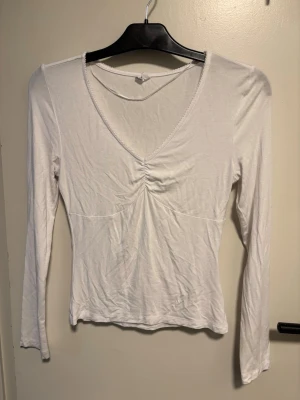 Vit långärmad topp med v-ringning - Säljer en enkel och stilren vit långärmad topp med v-ringning och rynkdetalj framtill. Toppen är figurnära och gjord i mjukt bomullsmaterial, perfekt att matcha med jeans eller kjol.