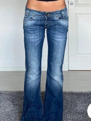 Blå bootcut jeans från Lee - Säljer ett par blå bootcut jeans från Lee med låg midja och klassiska fem fickor. Jeansen har snygga slitningar framtill och Lee-logga på bakfickan. Tillverkade i denim med en skön passform och utsvängda ben. Perfekta för en trendig och avslappnad look.