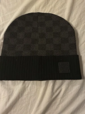 Svart och grå mössa - Snygg svart och grå mössa med diskret rutmönster och ribbad uppvikt kant. Märkeslogga på framsidan. Perfekt accessoar för att hålla värmen och lyfta din stil.