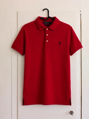 Polo Ralph Lauren röd piké – Custom Fit L (14–16) ❤️ - Klassisk röd piké från Polo Ralph Lauren i Custom Fit. Marinblå broderad Polo-logga på bröstet, tre knappar framtill och små slitsar längst ner. Tidlös modell som passar perfekt till jeans, chinos eller shorts. Storlek: L/G (14–16), ungdom Skick: Mycket fint skick – inga tydliga defekter syns på bilderna. Skriv gärna för mått eller fler bilder!