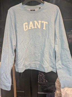 Ljusblå sweatshirt från GANT - Ljusblå sweatshirt från GANT med klassisk rund halsringning och stora vita bokstäver på bröstet. Tröjan har långa ärmar och ribbade muddar vid ärmslut och nederkant. Inga synliga fläckar eller defekter. Skapa en bundle för bättre pris