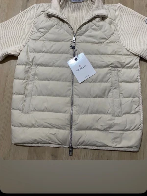 Vit dunväst från Moncler - Helt nya vit dunväst från Moncler med stickade detaljer och hög krage. Västen har dragkedja framtill, två sidofickor och quiltad design. Perfekt för lager-på-lager och ger en clean look. Materialet är en mix av dun och stickad bomull.