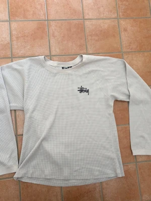 Vit långärmad topp från Stüssy - Vit långärmad topp från Stüssy med svart logga på bröstet och stor logga på ryggen. Tillverkad i ett ribbat material som ger en avslappnad streetwear-look. Perfekt till jeans eller cargobyxor för en chill vibe.