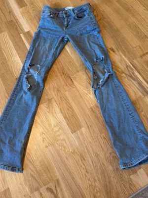 Blå slitna bootcut jeans från Zara - Säljer ett par blå bootcut jeans från Zara med stora slitningar över båda knäna. Jeansen har klassisk femficksmodell, hög midja och är tillverkade i denim. Perfekta för dig som gillar en avslappnad och trendig stil.