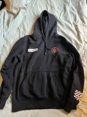 Svart Formula 1 hoodie med tryck - Svart hoodie med huva och känguruficka. Framsidan har Formula 1-logga och ett F1-märke, medan baksidan har stort Formula 1-tryck och en racerbilsbild. Vit rutdetalj på ena ärmen. Perfekt för motorsportfans!