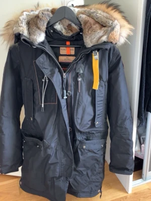 Svart parajumper jacka med pälsfodrad huva - Snygg svart parkas från Parajumpers i storlek S med stor pälsfodrad huva och gul detalj vid halsen. Jackan har flera fickor med dragkedja och knappar. Materialet är slitstarkt och jackan har en längre modell som skyddar mot kyla. Och är i mycket bra skick 