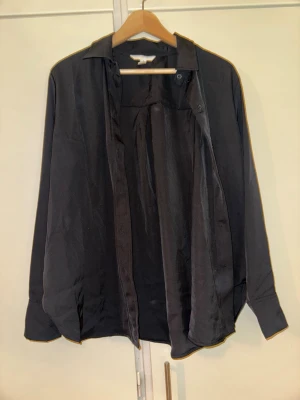 Svart skjorta från H&M, storlek M - Klassisk svart skjorta från H&M i storlek M. Skjortan har lång ärm, krage och knappar framtill. Tillverkad i ett mjukt och lätt material som ger en snygg och stilren look. Perfekt att styla lager på lager eller bära som den är.