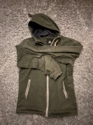 Lyle and Scott jacka  - Snygg olivgrön hoodiejacka med dragkedja framtill och två fickor med vita detaljer. Jackan har huva med mörkblått foder och elastiska muddar vid ärmsluten. Perfekt för en avslappnad streetstyle-look.