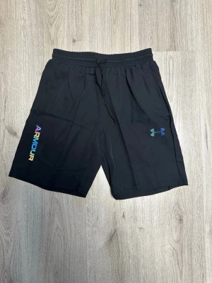 Svarta shorts från Under Armour - Snygga svarta shorts från Under Armour med regnbågsfärgad logga på ena benet och klassisk logotyp på andra. Shortsen har elastisk midja med snörning och är perfekta för träning eller chill. Tillverkade i lätt och bekvämt material.
