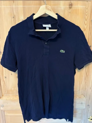 Marinblå pikétröja från Lacoste - Klassisk marinblå pikétröja från Lacoste med slim fit passform. Tröjan har korta ärmar, krage och knappar framtill samt den ikoniska gröna krokodilloggan på bröstet. Perfekt till jeans eller shorts för en clean och sportig look.