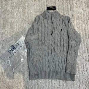 Grå kabelstickad tröja Polo Ralph Lauren - Snygg grå kabelstickad tröja från Polo Ralph Lauren med halv dragkedja och hög krage. Tröjan har ribbade muddar och den klassiska broderade loggan på bröstet. Perfekt för lager på lager och en stilren look.
