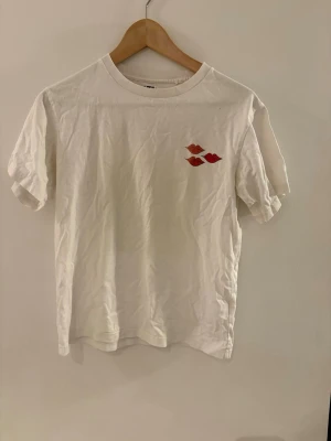 Vit t-shirt med tryck - Vit t-shirt med korta ärmar och broderade detaljer. Framtill finns små röda läppar och på ryggen syns en söt broderad kanin och björn med röda hjärtan ovanför. Perfekt för dig som gillar gulliga motiv och unika detaljer. Den är designad av konståret andy warhole och nypris är runt 600-700kr