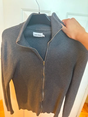 Grå ribbad ziptröja från East West - Snygg grå ribbstickad ziptröja från East West med hög krage och dragkedja hela vägen. Tröjan har långa ärmar och en klassisk, stilren look som passar till det mesta. Perfekt för lager på lager och enkel att matcha.
