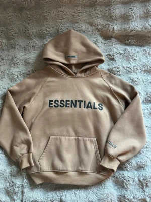 Essentials hoodie - Säljer en beige hoodie från Essentials med stor svart logga på bröstet och mindre tryck på ärmen. Hoodien har en klassisk känguruficka, ribbade muddar och en rymlig huva. Perfekt för en chill och avslappnad stil.