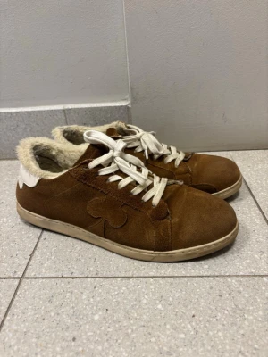 The Gallerist - St Moritz - Bruna sneakers i mocka med vita skosnören och vit detalj på hälen. Insidan är fodrad med mjukt, ljust fuskpälsfoder för extra komfort. Sidan har en diskret dekoration i samma färg som skon. Perfekta för dig som vill ha både stil och värme. Orginalkartongen finns ej kvar, men systrar skickas med. Finns en liten skada på båda skorna (se bild), men de är knappt märkbara. 