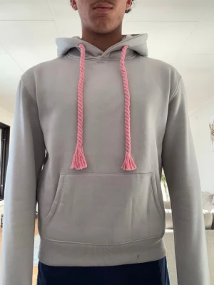 Syna world hoodie ljusgrå  - Snygg ljusgrå hoodie med huva och långa, tjocka rosa dragsnören. Klassisk känguruficka framtill och ribbade muddar vid ärmslut och nederkant. Perfekt för en avslappnad och trendig look.