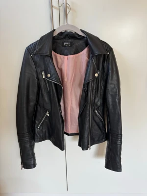 Svart skinnjacka från ONLY - Snygg svart skinnjacka från ONLY med silverfärgade dragkedjor och detaljer. Jackan har en klassisk bikerstil med bred krage och quiltade axelpartier. Insidan är fodrad med ljusrosa tyg. Perfekt för dig som vill ha en edgy look. Storlek 34💞
