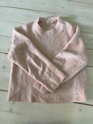 Ljusrosa stickad tröja Gina Tricot - Mysig ljusrosa stickad tröja från Gina Tricot i ullblandning. Tröjan har rund halsringning, ribbade muddar vid ärmslut och nederkant samt en avslappnad passform. Perfekt för kyliga dagar och enkel att styla med jeans eller kjol.