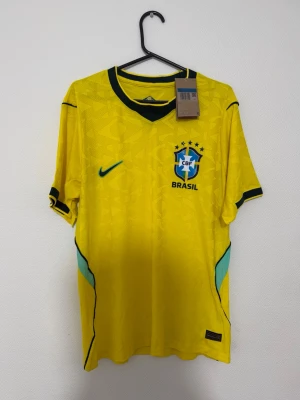 Brasilien matchtröja gul Nike - Gul Brasilien matchtröja från Nike med gröna detaljer och svart krage. Tröjan har CBF-emblemet och Nike-logga på bröstet samt diskret mönster i tyget. Perfekt för fotboll och sportiga aktiviteter.