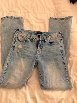 Ljusblå True Religion jeans - Säljer ett par ljusblå jeans från True Religion med klassiska röda sömmar och detaljerade bakfickor. Jeansen har en liten slitning framtill och är tillverkade i denim. Rak passform och femficksmodell med knappgylf.