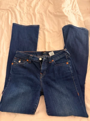 True Religion blå jeans med kontrastsömmar - Säljer ett par blå jeans från True Religion med ikoniska kontrastsömmar och snygga ficklock bak med guldknappar. Klassisk femficksmodell i mjuk denim med raka ben och normal midja. Perfekta för dig som gillar streetwear och vill sticka ut.