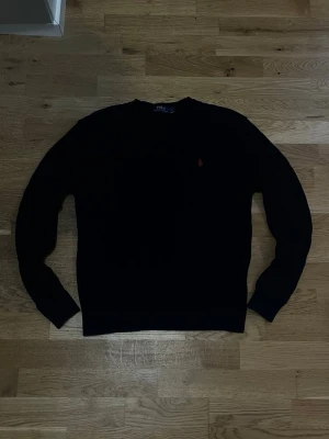 Svart stickad tröja med röd logga - Klassisk svart stickad tröja med rund hals och lång ärm. Tröjan har en liten röd logga broderad på bröstet och är enkel men stilren. Passar perfekt till jeans eller över en skjorta för en clean look.