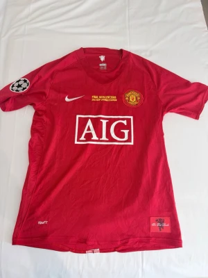 Manchester United Ronaldo matchtröja - Säljer en röd Manchester United matchtröja från Nike med AIG som sponsor, Champions League-märke på ärmen och Ronaldo 7 på ryggen. Tröjan har klubbmärke och finalbrodyr på bröstet samt vita detaljer. Perfekt för dig som älskar fotboll och vill ha en ikonisk supportertröja.