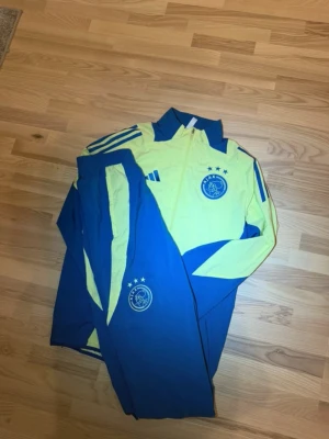 Ajax träningsdress blå/gul Adidas - Säljer en Ajax träningsdress från Adidas i blått och gult. Setet består av en jacka med dragkedja och matchande byxor, båda med Ajax-logga och Adidas ränder på axlar och ben. Perfekt för fotbollsträning eller chill. Materialet är syntetiskt och lätt.