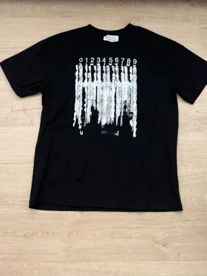 Svart t-shirt med tryck från Maison Margiela - Svart t-shirt från Maison Margiela med ett unikt vitt tryck framtill som ser ut som rinnande siffror. Klassisk passform och rund hals. Perfekt för dig som gillar statement-plagg och streetwear vibes.
