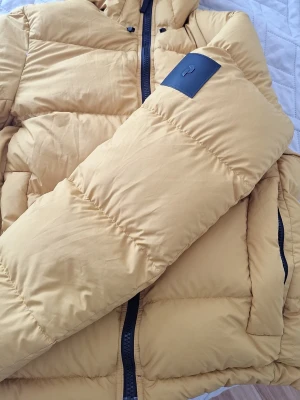 Gul pufferjacka från Peak Performance - Säljer en snygg gul pufferjacka från Peak Performance med huva och svarta detaljer. Jackan har en klassisk quiltad design, dragkedja framtill och Peak Performance-logga på ärmen. Perfekt för kalla vinterdagar och riktigt skön att bära.
