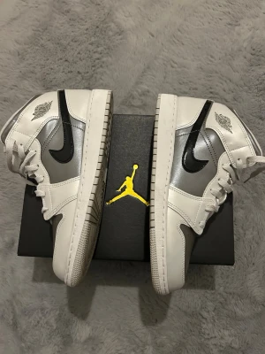 Air Jordan 1 Mid Silver/White/Black - Snygga Air Jordan 1 Mid sneakers i silver, vitt och svart. Skorna har klassisk svart swoosh, vita paneler och silverfärgade detaljer. Tillverkade i läder med ikonisk Jordan-logga på sidan. Perfekta för dig som vill sticka ut med stilrena och sportiga sneakers.