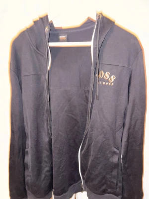 Svart hoodie från BOSS med dragkedja - Säljer en svart hoodie från BOSS med dragkedja framtill och huva med snörning. BOSS-logga i guld på bröstet och detaljerad text längs huvan. Perfekt för en chill och stilren look. Sitter som M