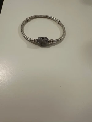 Pandora armband i silver  - Snyggt Pandora armband i silverfärg med en glittrande hjärtformad pärla i mitten. Armbandet har en stilren och modern design med detaljerade mönster på pärlan. Perfekt accessoar för att lyfta din outfit.