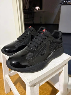 Svarta sneakers med glansig finish - Snygga helsvarta sneakers med glansiga partier och meshdetaljer på sidorna. Skorna har svarta skosnören, låg profil och en chunky sula. Perfekta för dig som gillar en clean och modern look. Små röda detaljer på plösen ger en extra touch.