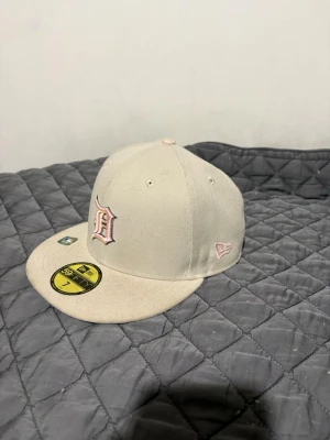 Beige New Era keps med brodyr - Snygg beige keps från New Era med rosa broderad D-logga framtill och cool tigerbrodyr med röda rosor på sidan. Klassisk flat brim och broderade ventilationshål. Passar perfekt för dig som vill sticka ut med stilrena detaljer.