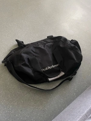 Svart duffelväska Peak Performance - Snygg svart duffelväska från Peak Performance med axelrem och två bärhandtag. Väskan har dragkedja upptill och logga i vitt på sidan. Perfekt för gymmet eller resan, tillverkad i slitstarkt tygmaterial.