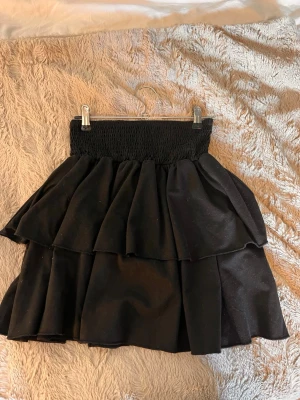 Svart volangkjol med resårmidja - Snygg svart kortkjol med dubbla volanger och bred smockresår i midjan. Kjolen har en luftig och lekfull look som passar perfekt till olika stilar. Materialet känns mjukt och följsamt.