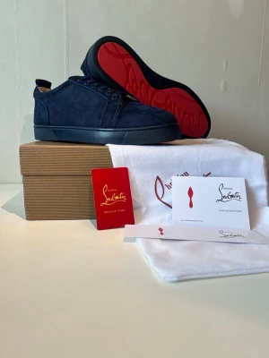 Louboutin skor - Snygga marinblå sneakers från Christian Louboutin i mocka med den ikoniska röda sulan. Skorna har klassisk låg profil, rund tå och matchande blå snörning. Perfekta för dig som vill ha en exklusiv och stilren look.