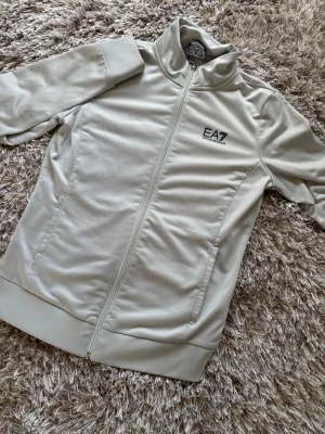 Ljusgrå ziptröja EA7 Emporio Armani - Snygg ljusgrå ziptröja från EA7 Emporio Armani med hög krage och dragkedja framtill. Tröjan har två fickor på sidorna och svart logga på bröstet. Tillverkad i ett mjukt och stretchigt material som passar perfekt till sport eller chill.
