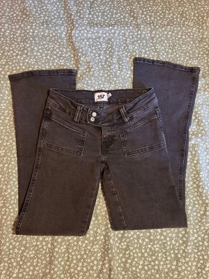 Svarta bootcut jeans med låg midja - Snygga svarta jeans med bootcut och låg midja. Jeansen har dubbla knappar i midjan, breda bälteshällor och snygga framfickor. Perfekta för dig som gillar en 2000s vibe och vill ha ett par jeans som sticker ut lite extra.