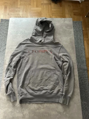Grå hoodie från C.P. Company - Säljer en grå hoodie från C.P. Company med tryck i rött framtill och ett stort grafiskt tryck på ryggen. Hoodien har känguruficka och ribbade muddar vid ärmar och nederkant. Perfekt för en avslappnad streetwear-look.