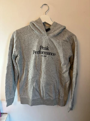 Peak Performance hoodie - Snygg grå hoodie från Peak Performance med svart logga och text på bröstet. Klassisk känguruficka framtill och ribbade muddar vid ärmar och nederkant. Perfekt för en avslappnad och sportig stil. Tillverkad i mjukt bomullsmaterial och har huva.