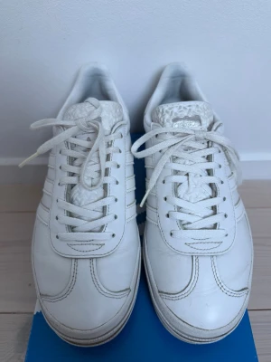 Adidas Gazelle Bold W vita sneakers - Säljer ett par vita Adidas Gazelle Bold W sneakers med klassisk design och tjockare sula. Skorna har vita läderpaneler, vita snören och diskreta Adidas-detaljer på plösen. 