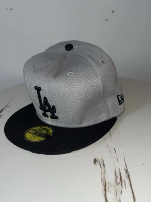 Grå LA New Era 59FIFTY keps - Snygg grå keps från New Era 59FIFTY med svart skärm och LA-logga framtill. Klassisk snapback-modell med broderade detaljer och svart knapp på toppen. Perfekt för dig som gillar streetwear och sportig stil.