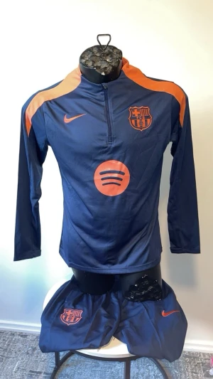 Barcelona träningsset blå/orange Nike - Snyggt träningsset från FC Barcelona i marinblått med orange detaljer. Setet består av långärmad tröja med halv dragkedja, shorts och långa träningsbyxor. Alla delar har klubbmärke och Nike-logga samt sponsortryck på tröjan. Perfekt för fotbollsträning eller chill.