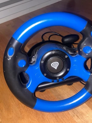 Genesis ratt och pedaler till spelkonsol - Säljer en blå och svart ratt från Genesis med tillhörande pedaler. Ratten har flera knappar, inklusive LB och X/Y, och är tillverkad i plast. Perfekt för racing- och bilspel på konsol eller PC.
