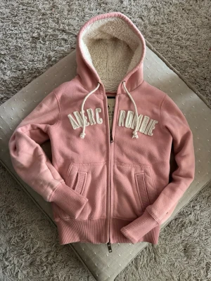 Rosa Abercrombie & Fitch Hoodie med teddyfoder - Mysig rosa hoodie från Abercrombie med dragkedja, fickor och vit brodyr på bröstet. Hoodien är fodrad med fluffigt teddyfoder och har vita dragsnören. Perfekt för chill dagar eller när du vill vara extra bekväm.