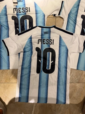 Argentina Hemmaträja Messi #10 - S - Säljer en Argentina matchtröja med Messi och nummer 10 på ryggen. Tröjan har klassiska blå och vita vertikala ränder, svarta detaljer på axlar och ärmslut samt officiella märken på bröstet. Perfekt för dig som älskar fotboll och vill stötta ditt favoritlag.