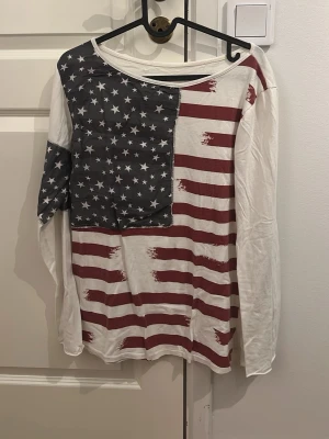 Vit långärmad topp med USA-flagga - Vit långärmad topp med tryck av amerikanska flaggan framtill. Toppen har rund halsringning och är tillverkad i mjuk bomull. Mönstret består av röda ränder och vita stjärnor på blå botten. Perfekt för dig som gillar statement-plagg.