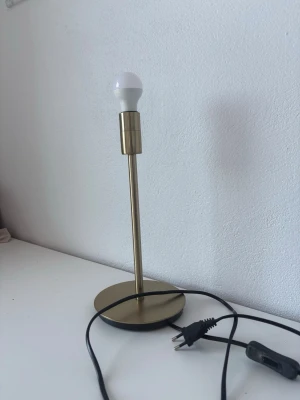 Bordslampa i mässing från IKEA - Snygg bordslampa i mässingsfärgad metall från IKEA. Lampan har en rund fot, smal stång och medföljande LED-lampa. Passar perfekt på skrivbordet eller nattduksbordet. Strömkabel med stickkontakt ingår.